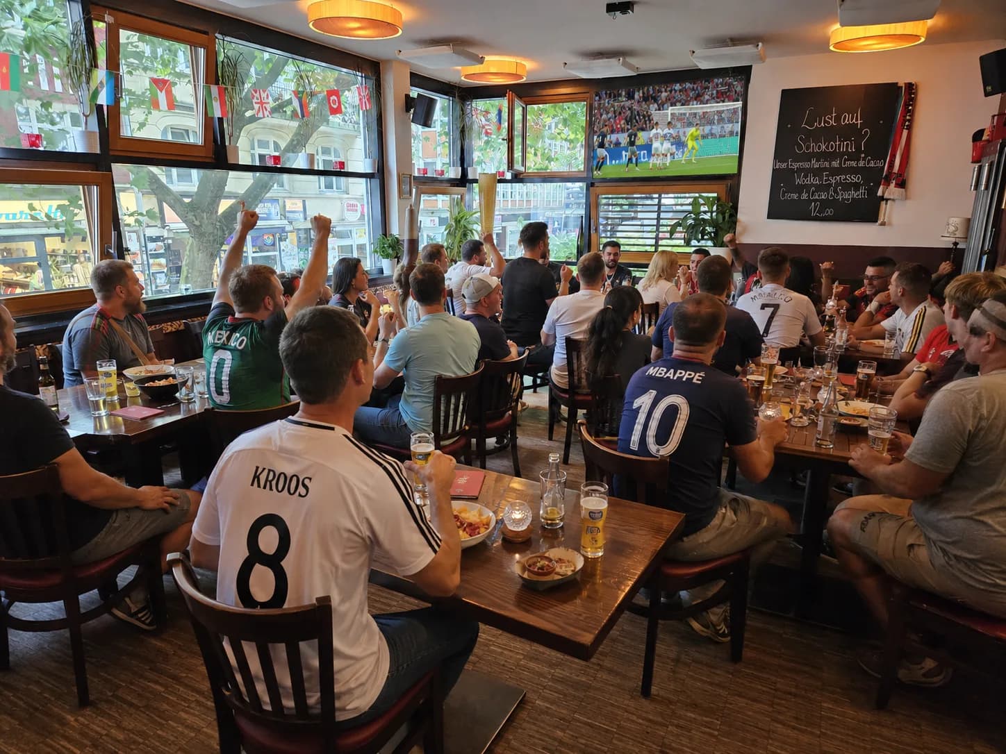 Tagsüber gefüllter Südhang in Hamburg-Sternschanze mit Gästen, Fenstern zur Schanze und Fußball-Leinwand